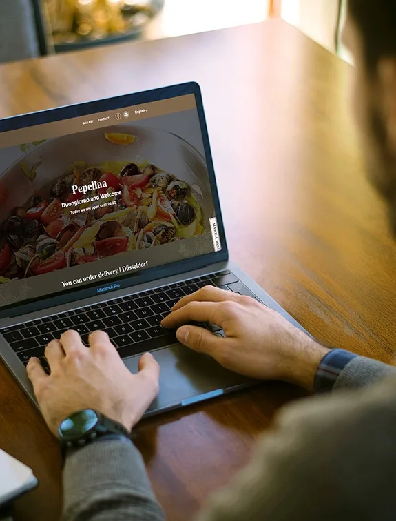 Osoba pracuje na notebooku s otevřenou webovou stránkou restaurace; viditelné pokrmy a možnosti objednání.