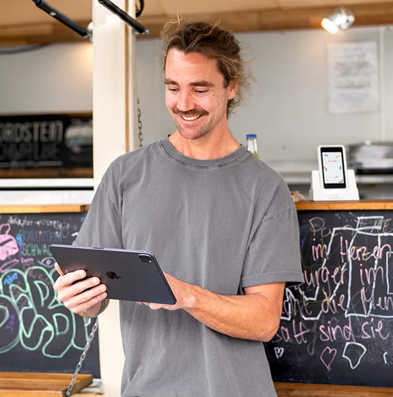 Muž stojí před food truckem a používá tablet; v pozadí je tabule s menu.