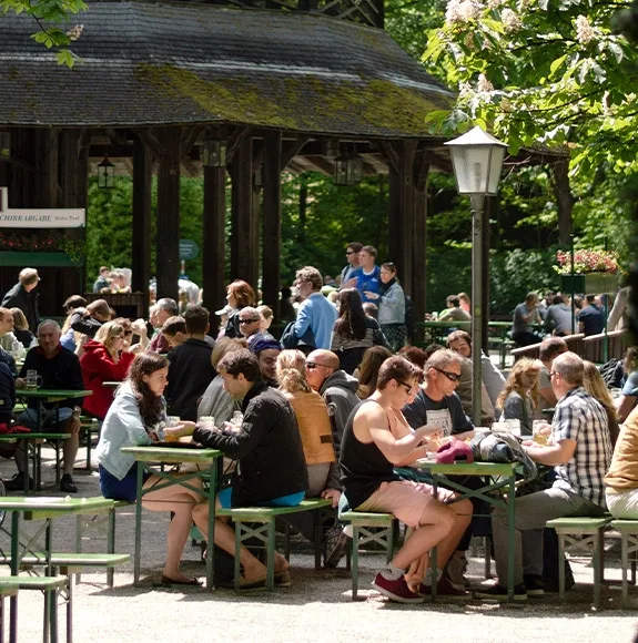 People sitting at green wooden tables in a shady beer garden enjoying drinks outdoors.