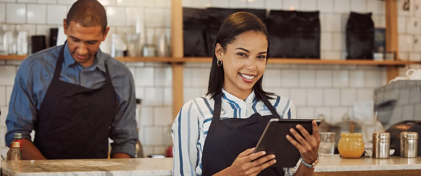 Two café employees: One employee is holding a tablet and smiling, while her colleague is working in the background.