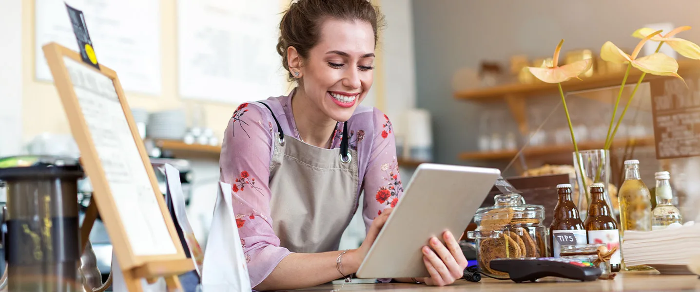 Café employee stands behind the counter and checks a tablet while managing orders.