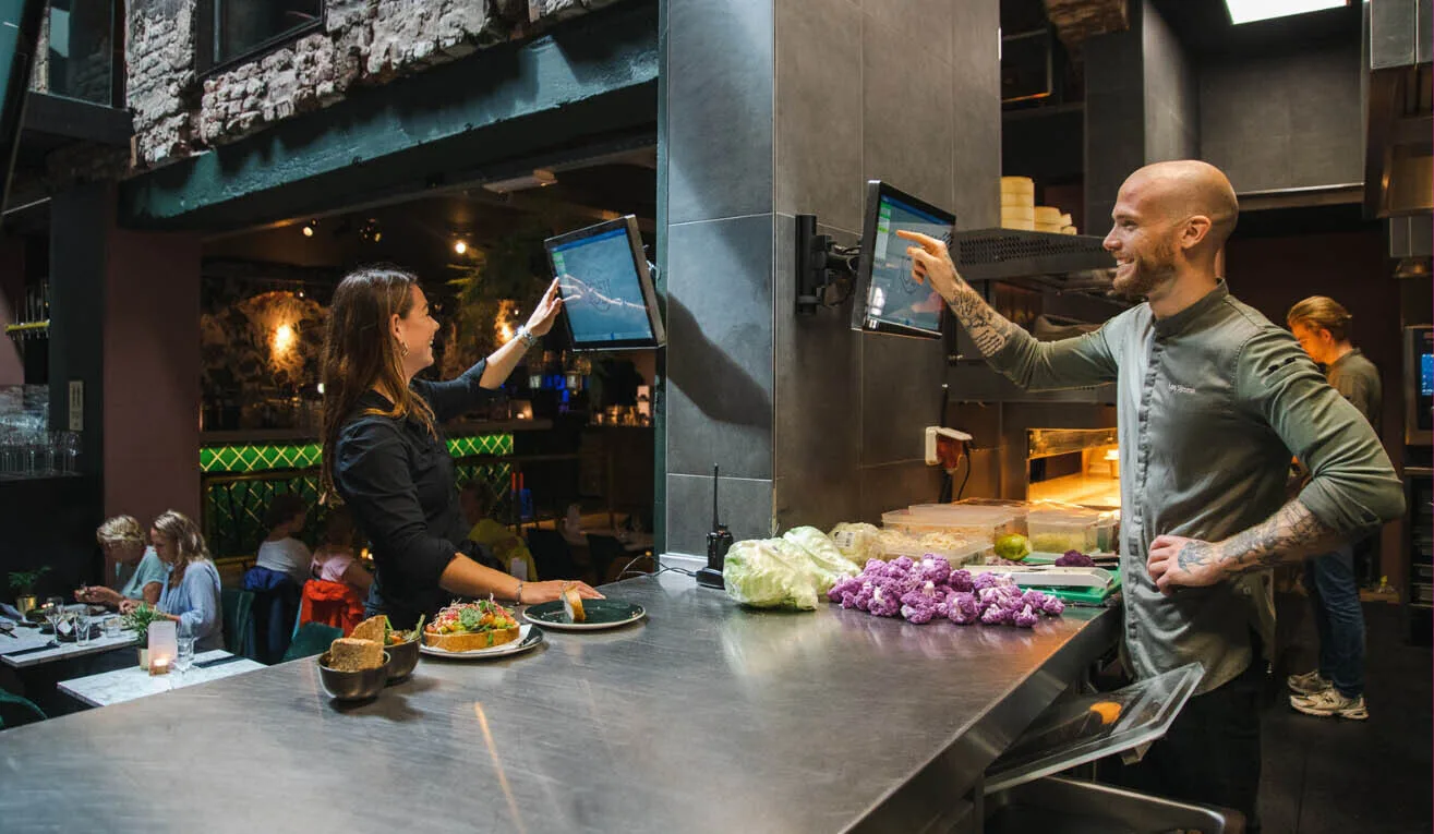 Restaurant staff using digital kitchen display screens to manage orders between the kitchen and service area.