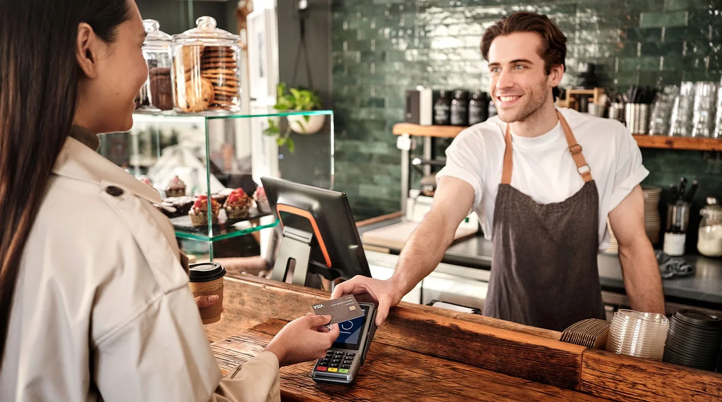 Customer with coffee cup pays contactless with card in café; café employee hands over payment terminal across the counter.