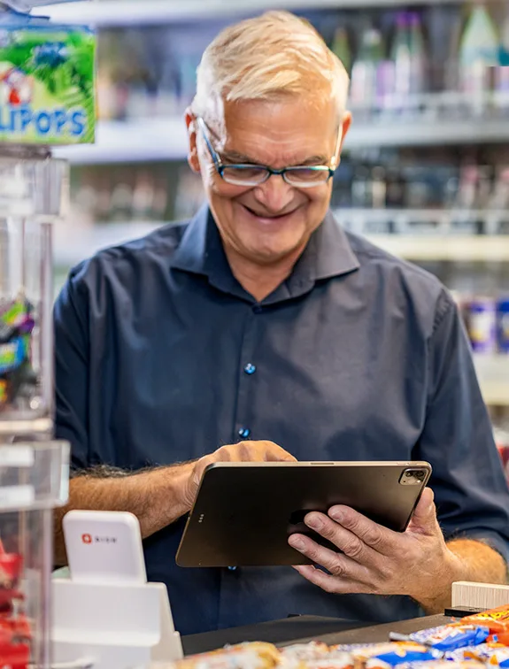 Elderly man stands at the checkout with a tablet; next to him is a white card reader.