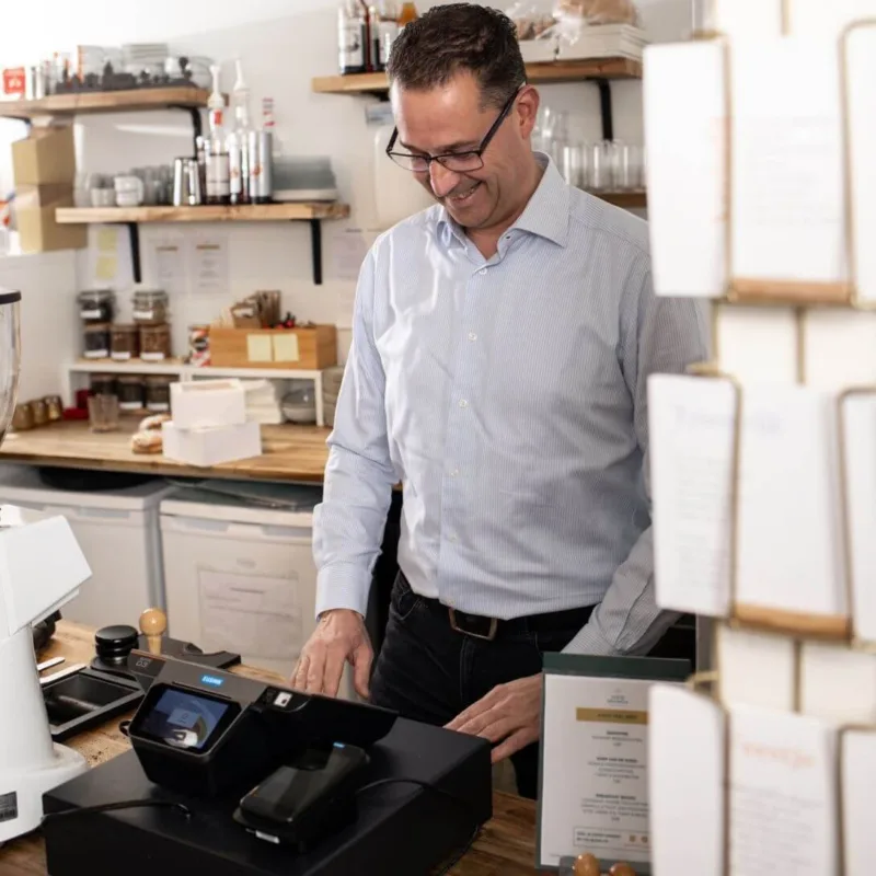 A man in a blue shirt stands behind a cafe counter, smiling while using a touchscreen payment terminal.