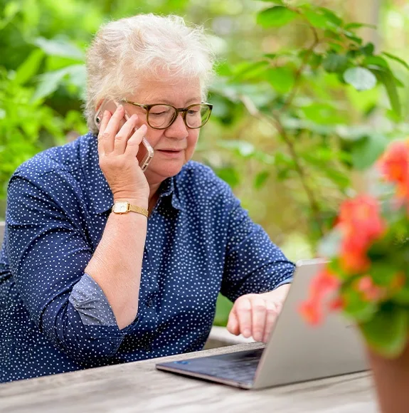An elderly woman is talking on the phone and working on a laptop at the same time in a beer garden.