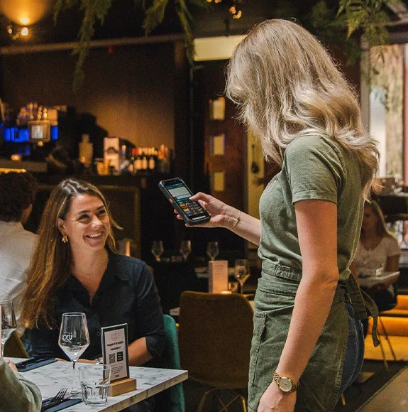 Waitress holding a mobile payment terminal while a guest sits at a table laughing.