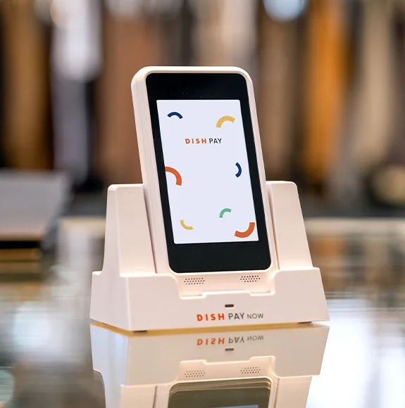 Small white card reader with the inscription “DISH Pay Now” stands on a table. The screen shows the logo and colorful graphic elements.