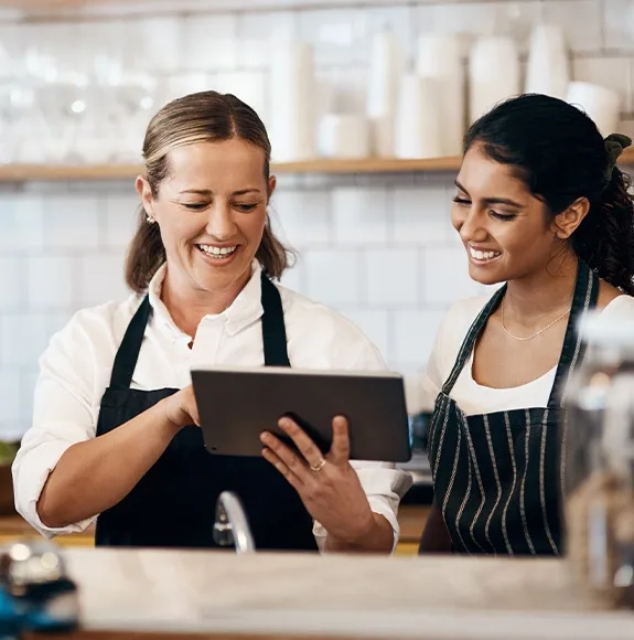A café owner and her employee look together at a tablet behind the counter and discuss something.