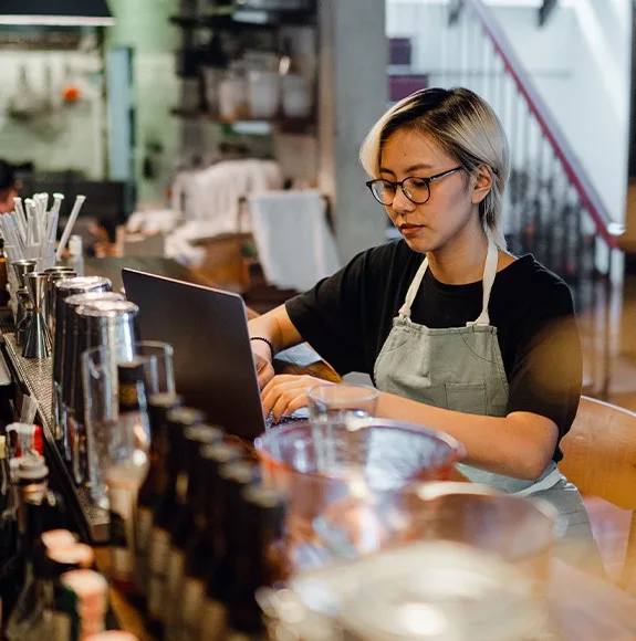 A waitress wearing an apron works intently on a laptop behind the bar of a restaurant.