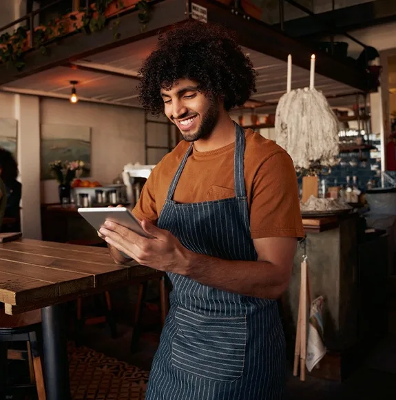 A smiling waiter wearing an apron checks reservations on a tablet while standing at a counter.