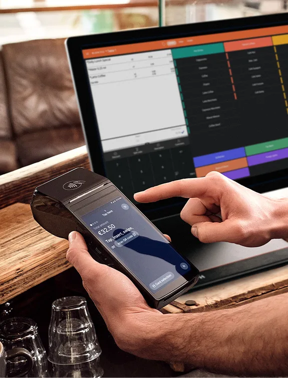 A waiter is entering a sum with his finger on a mobile digital payment terminal next to the POS screen.