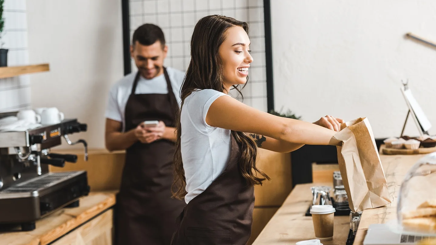 Two café employees prepare orders, one person packs a paper bag, while someone else looks at their smartphone in the background.