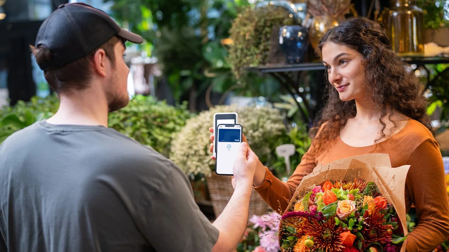 Florist holds a bouquet while a customer makes a contactless payment with their smartphone at a white card reader.
