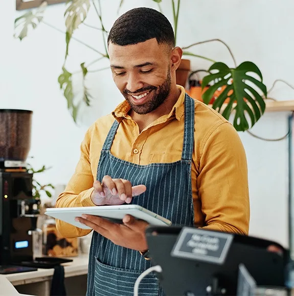 Un employé de café en chemise jaune et tablier rayé utilise une tablette au comptoir et sourit.