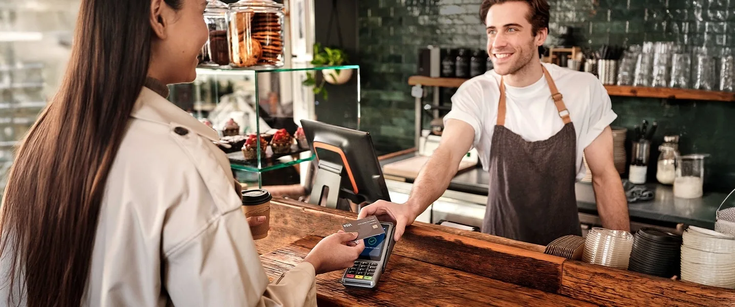 Un barista tend un terminal de paiement à une cliente au comptoir du café, où elle paie avec une carte de crédit Visa.