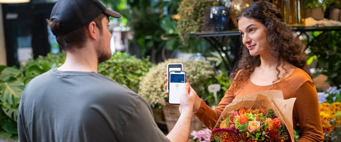 Un fleuriste tient un bouquet tandis qu'un client effectue un paiement sans contact avec son smartphone sur un lecteur de carte blanc.