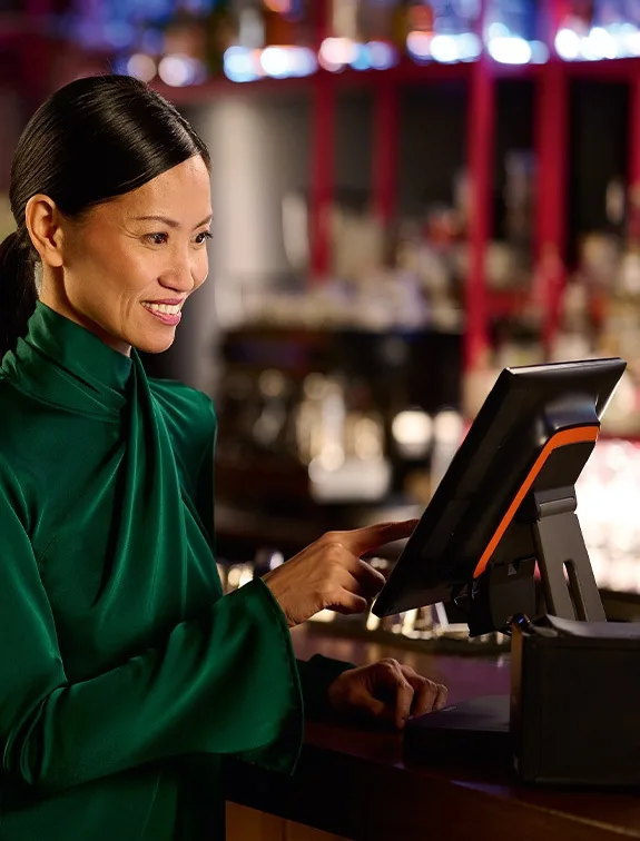 Une serveuse en tenue verte utilise un système de point de vente au bar et sourit.