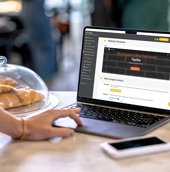 Une personne travaille sur un ordinateur portable avec un modèle de site web ouvert dans le café ; des croissants sous une cloche en verre à côté d'elle.