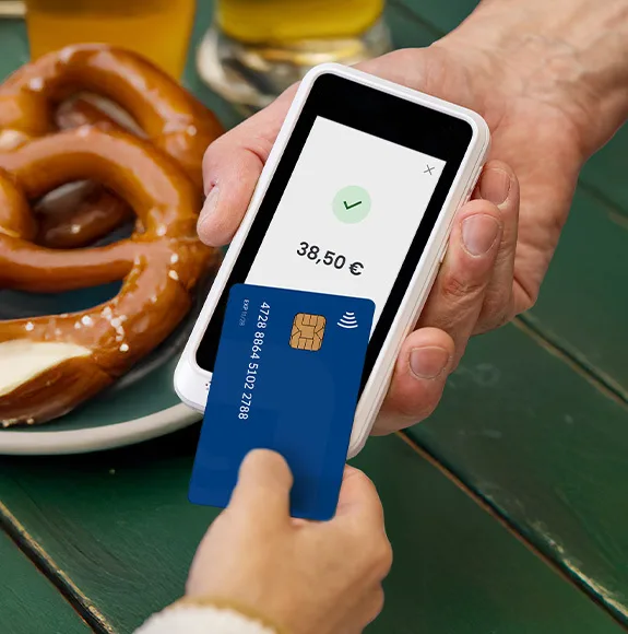 Kontaktlose Zahlung an einem Bezahlterminal neben einem Teller mit Brezeln und Getränken.