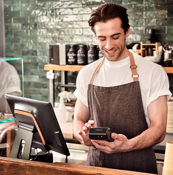 Café-Mitarbeiter mit Schürze bedient ein Bezahlterminal hinter der Theke; Kaffeemaschinen im Hintergrund.