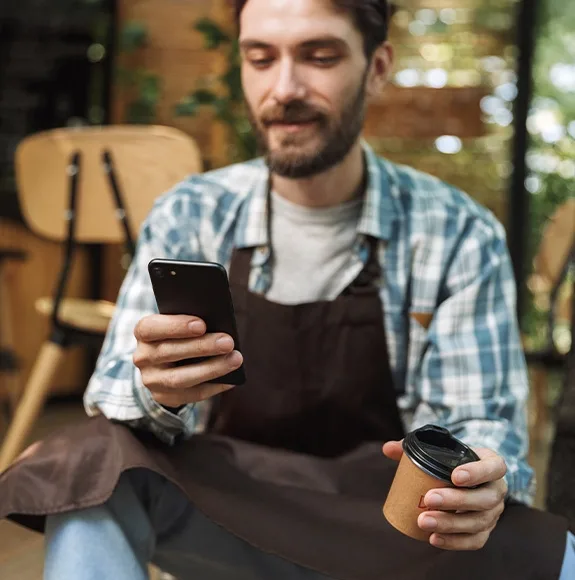 Kellner in kariertem Hemd und Schürze sitzt im Café, hält Smartphone in der einen und einen Kaffeebecher in der anderen Hand.