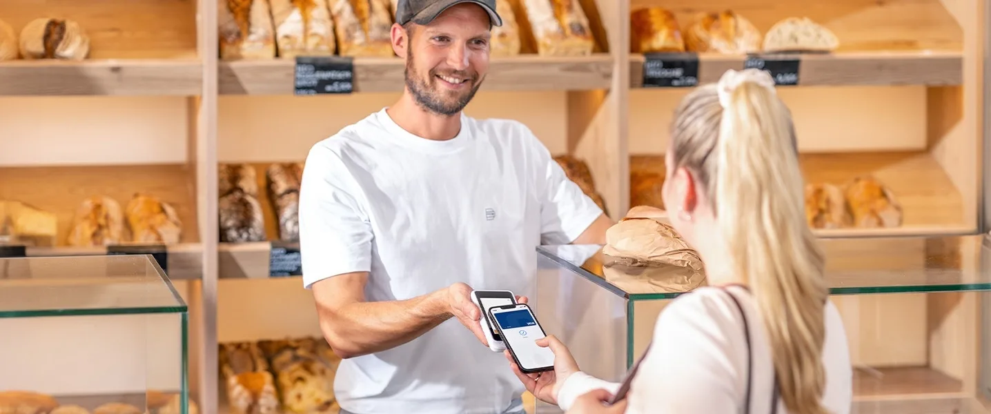 Ein Barista reicht einer Kundin an der Cafétheke ein Bezahlterminal, an dem sie mit einer Visa-Kreditkarte bezahlt.