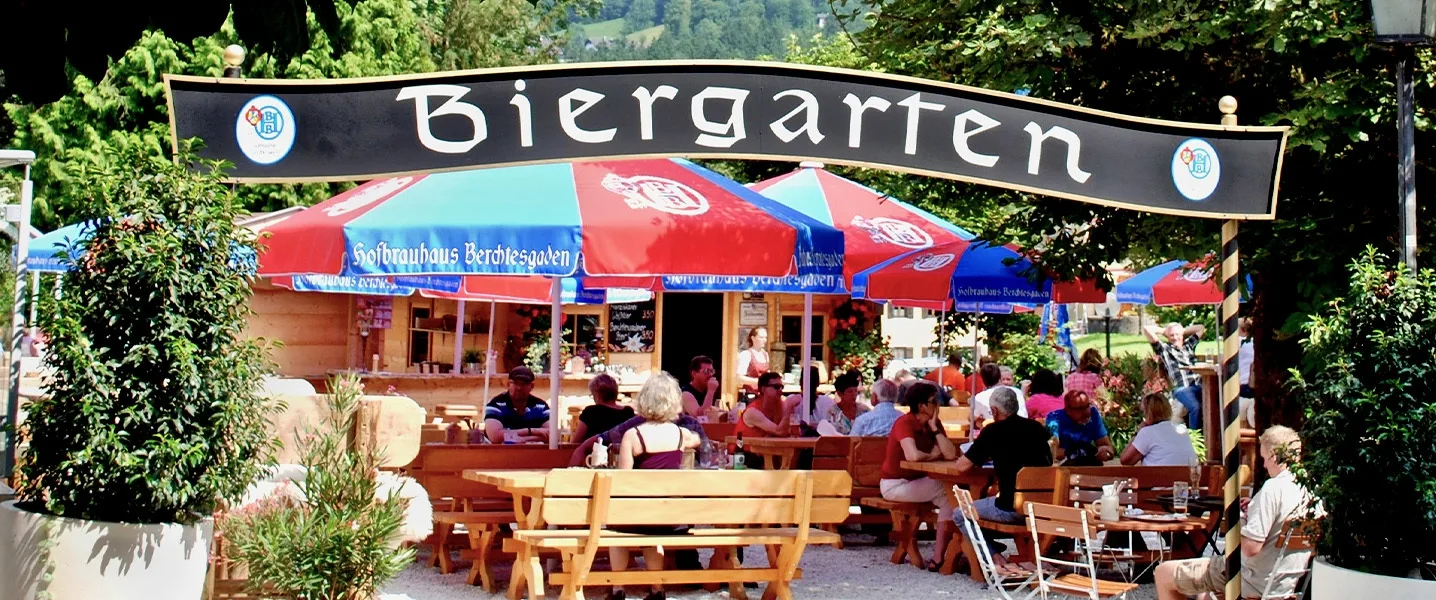 Eingang zu einem Biergarten mit großen Sonnenschirmen und Gästen an Holztischen.
