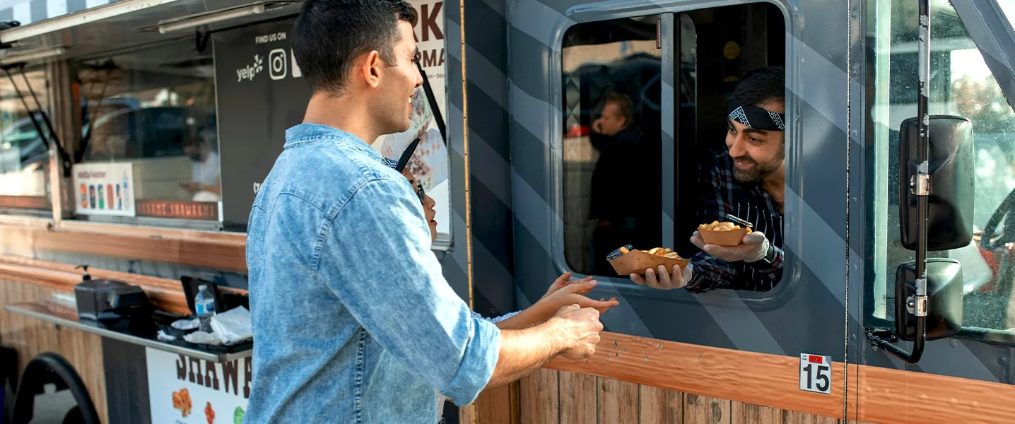 Verkäufer reicht durch das Fenster eines Foodtrucks zwei Schalen mit Essen an einen Kunden.