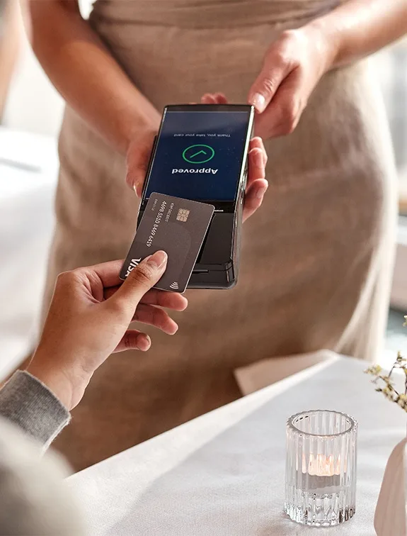 Nahaufnahme einer Hand, die eine Kreditkarte an ein mobiles Bezahlterminal hält und eine Zahlung durchführt.