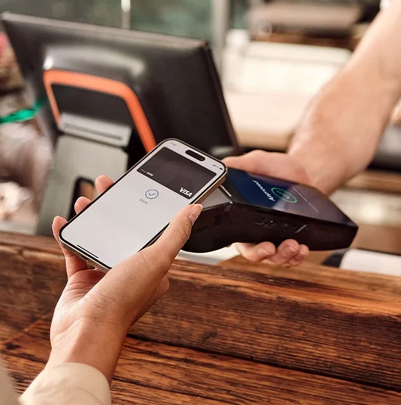 Person bezahlt kontaktlos mit dem Smartphone an einem Bezahlterminal auf einer Cafétheke.
