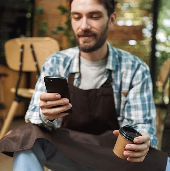 Cameriere con camicia a quadri e grembiule seduto nel caffè, tiene uno smartphone in una mano e una tazza di caffè nell'altra.