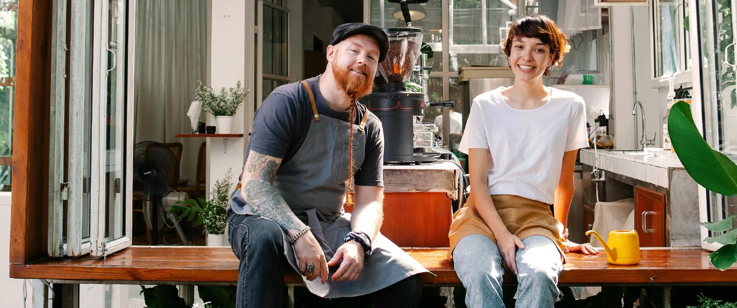 Due giovani dipendenti di un caffè siedono sorridenti su una panchina davanti a una finestra luminosa.