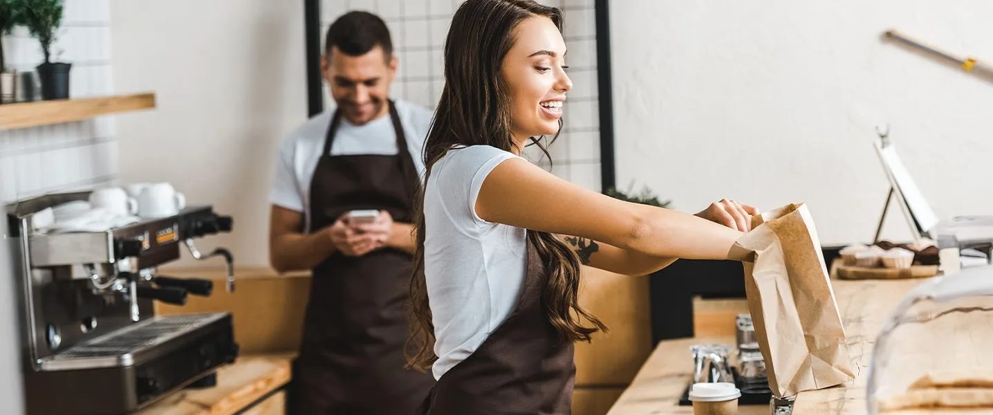 Due dipendenti del bar preparano gli ordini, una persona riempie un sacchetto di carta, mentre qualcun altro guarda il proprio smartphone sullo sfondo.