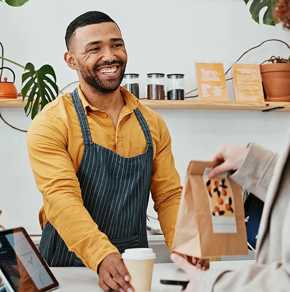 Un dipendente di un caffè consegna una borsa di carta e una tazza di caffè a un ospite con un sorriso amichevole.