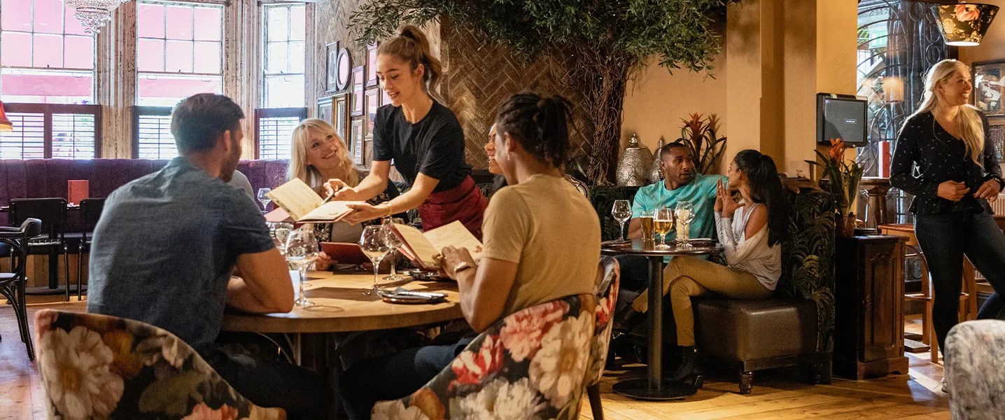 Kelnerka obsługuje gości w stylowo urządzonym restauracji; ludzie przeglądają menu.