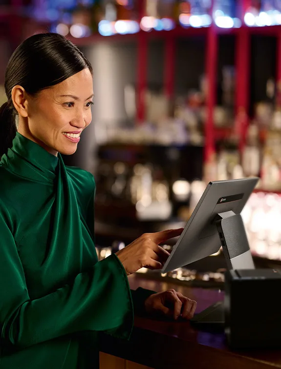 Kelnerka w zielonym stroju obsługuje system POS przy barze i uśmiecha się.