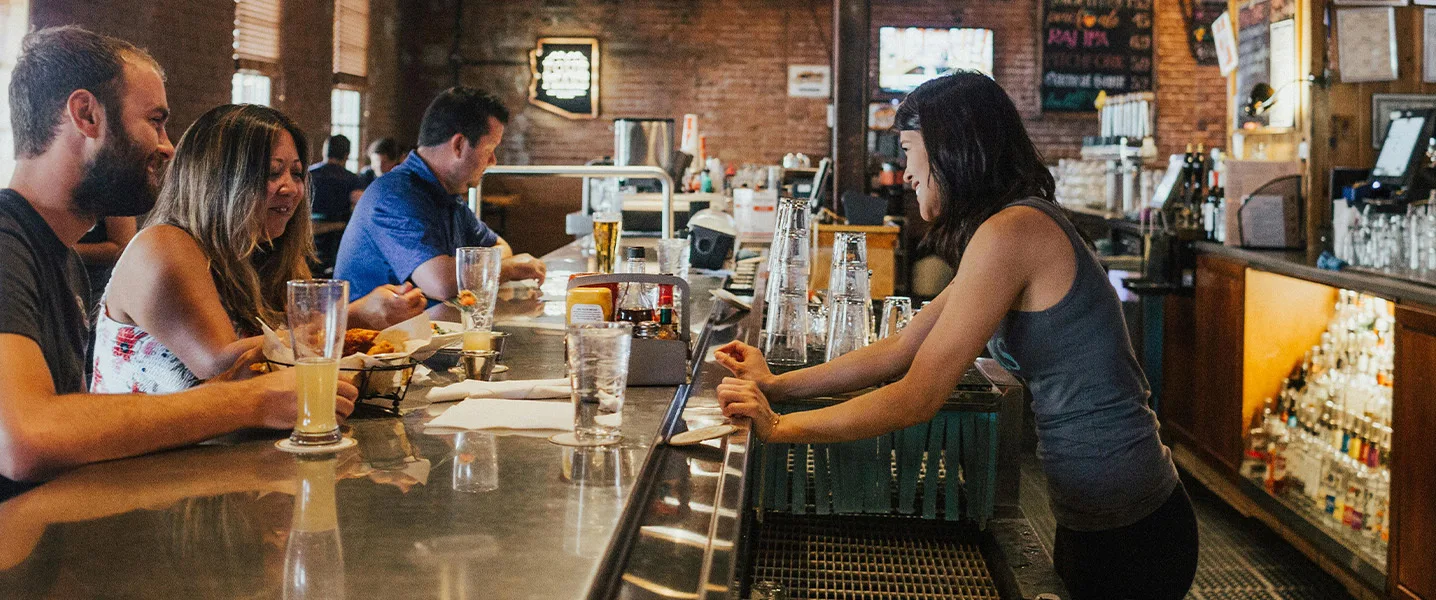 Empregada de bar inclina-se sobre o balcão e conversa com clientes que estão a comer e a beber.