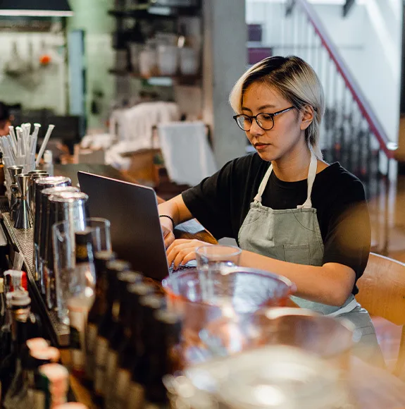 Uma empregada de avental trabalha concentrada num portátil atrás do bar de um restaurante.