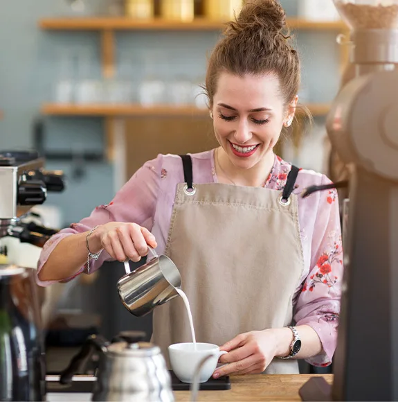 Zamestnanec kaviarne nalieva mlieko do šálky kávy a usmieva sa pri espresso stroji.