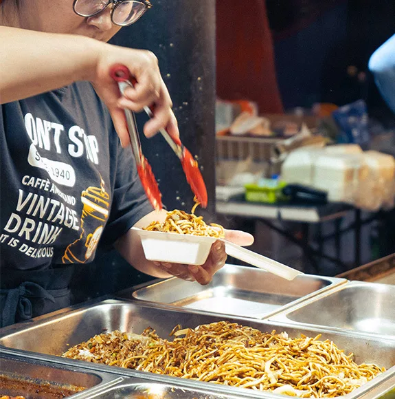 Persona sirve fideos fritos con pinzas en un recipiente desechable blanco en un puesto de comida.