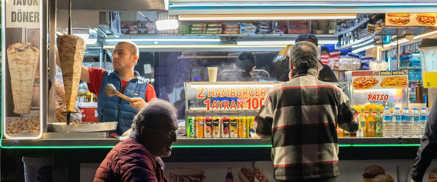 Bar de snacks con clientes; vendedor corta carne de un pincho de kebab, tablero de menú iluminado en el fondo.