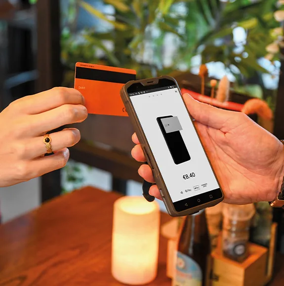 La mano de una mujer sostiene una tarjeta de crédito naranja contra la parte trasera de un terminal de pago sin contacto de DISH POS, en cuya pantalla se muestra el importe a pagar.